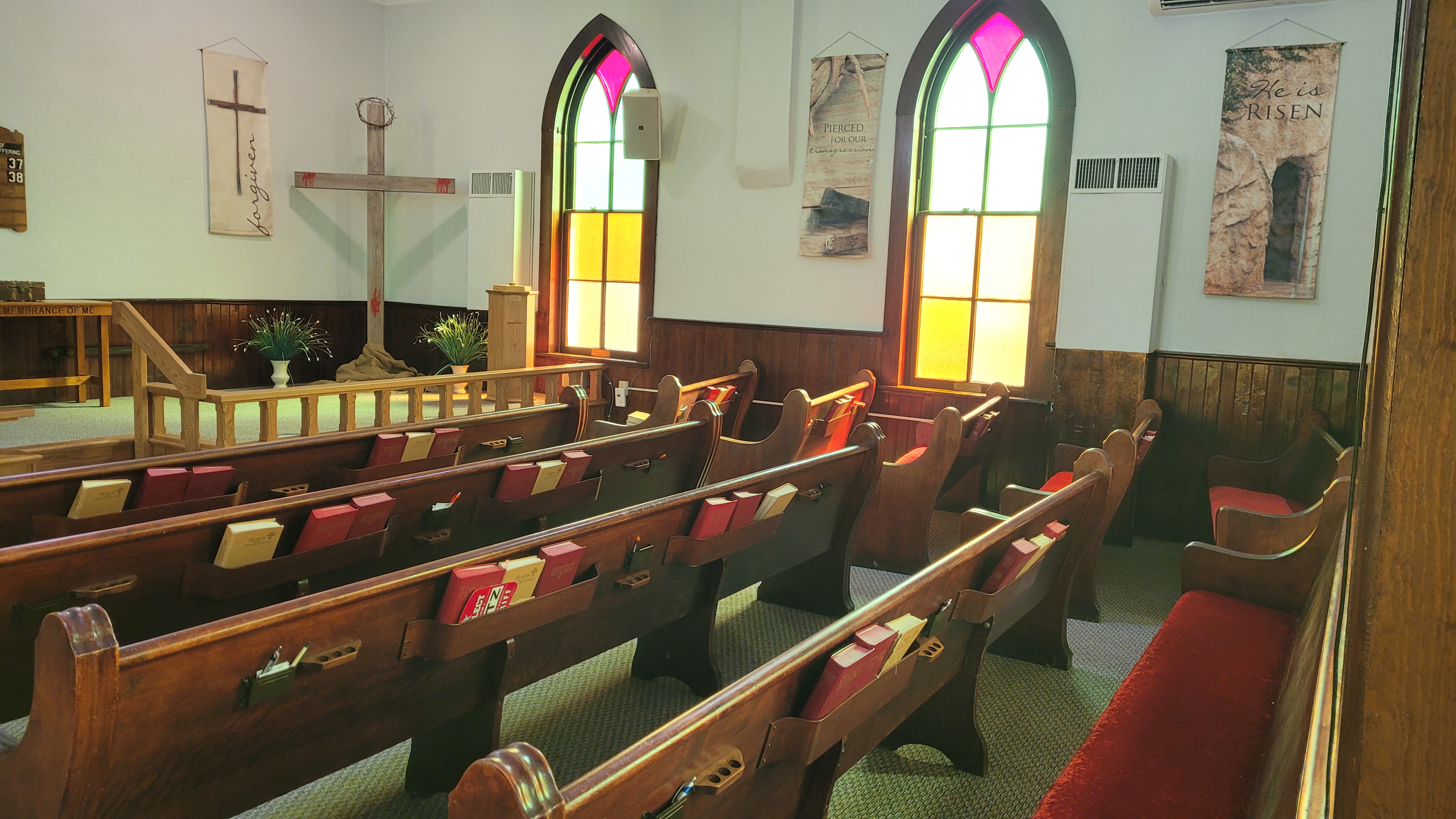 Two arched stained glass windows glowing gold and pink beside a rough wooden cross draped with cloth and a crown of thorns.